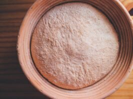 Imagem com a foto de cima de uma bacia de barro sobre uma tábua. Dentro dela, uma massa de pão rústica em formato de bola.