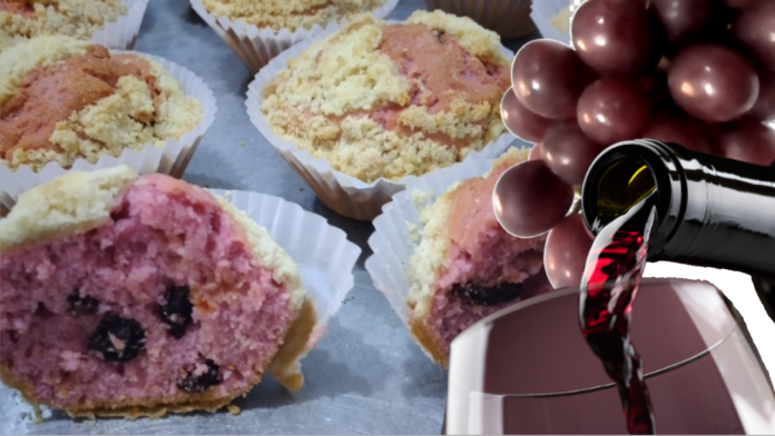 Imagem com a foto de cinco muffins, um deles cortado ao meio, mostrando a massa de cor de uva, com algumas uvas passas no recheio. Na frente, parte de uma taça aparece, com a ponta de uma garrafa servindo vinho, e algumas uvas frescas.