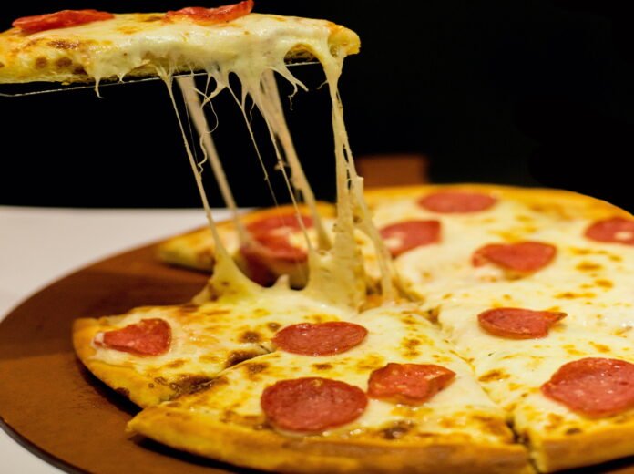 Pizza com muito queijo e fatias de calabresa, sobre uma tábua de madeira redonda. Undo preto e uma espátula levantando uma fatia da pizza.