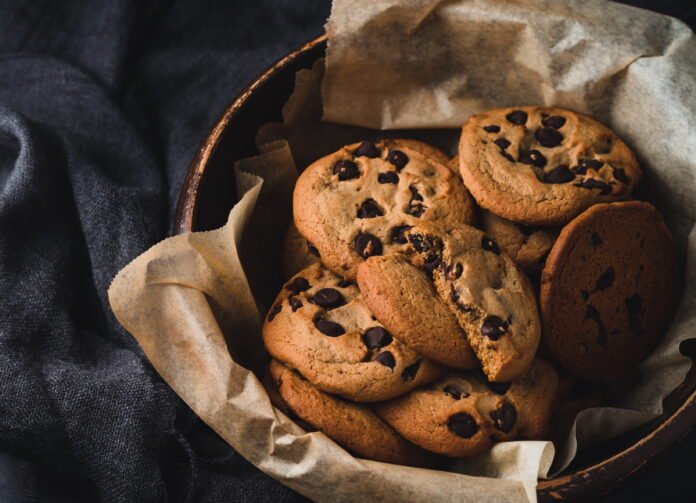 Foto de uma bacia de metal forrada com um papel e cheida de biscoistos redondos com gotas de chocolates. A bacia está sobre um tecido jeans.