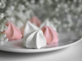 Foto de um prato de louça branca, dentro dele cinco suspiros doces. Três brancos e dois cor de rosas. A volta no prato, arranjos com pequeninas flores brancas gipsófilas.