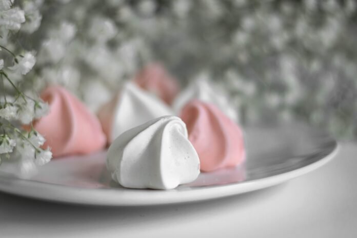 Foto de um prato de louça branca, dentro dele cinco suspiros doces. Três brancos e dois cor de rosas. A volta no prato, arranjos com pequeninas flores brancas gipsófilas.