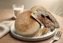 X-Assado à Portuguesa prato de porcelana branca com dois pães redondos, um está corrado pela metade com recheio de frios aparecendo. O prato está sobre uma mesa juntamente com um pano branco, um garfo e um copo com leite.