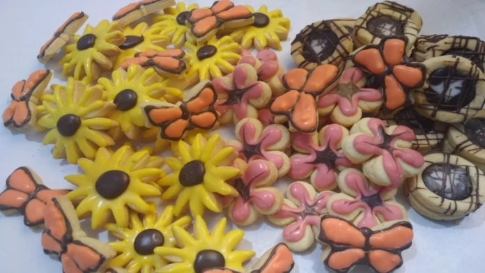 Várias bolachas artesanais com formatos variados. Margaridas amarelas, borboletas laranjas, flores de cinco pétalas rosas e biscoitos com formato circular com recheio de chocolate.