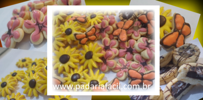 Várias bolachas artesanais com formatos variados. Margaridas amarelas, borboletas laranjas, flores de cinco pétalas rosas e biscoitos com formato circular com recheio de chocolate.