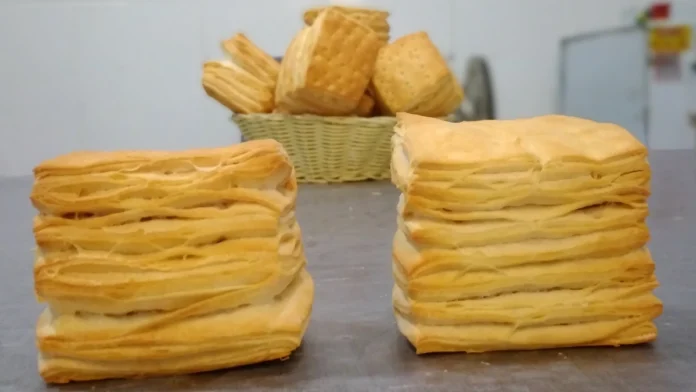 Dois pães quadrados e folhados à rente de uma cesta de vime com mais pães iguais.