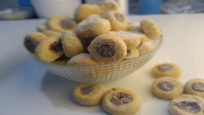 Imagem em formato de pergaminho. Nele, a foto de uma vasilha de vidro, cheia de pequenos biscoitos doces com um pingo de goiabada no meio, polvilhados com açúcar.