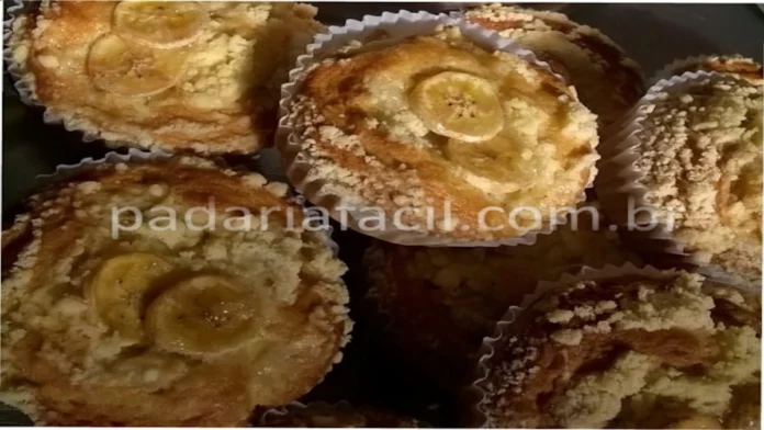 Vista superior de um amontoado de mini bolinhos de banana dentro de forminhas de papel. A superfície dos bolos de banana é coberta com farofa doce e algumas rodelas de banana.