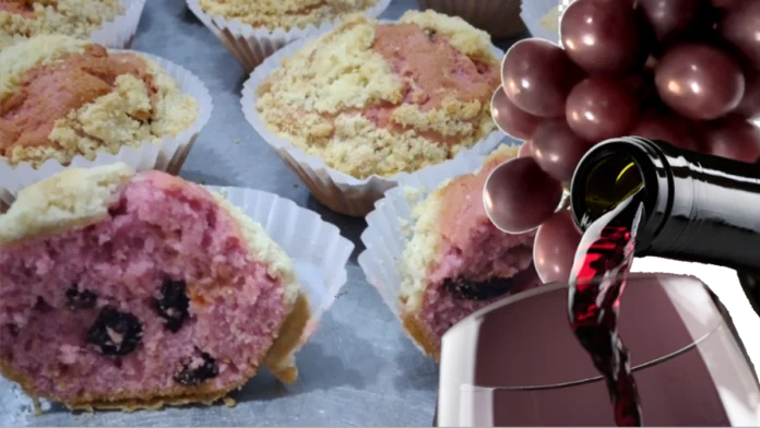 Imagem com a foto de cinco muffins, um deles cortado ao meio, mostrando a massa de cor de uva, com algumas uvas passas no recheio. Na frente, parte de uma taça aparece, com a ponta de uma garrafa servindo vinho, e algumas uvas frescas.