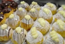 Sonho Assado Três bandejas brancas com diferentes tipos de pães. A primeira bandeja contém pães redondos cobertos com coco e creme. A segunda bandeja tem pães cobertos com ganache branco, riscos de chocolate preto e gotas de creme. A terceira bandeja apresenta pães cobertos com chocolate preto e gotas de creme de chocolate.