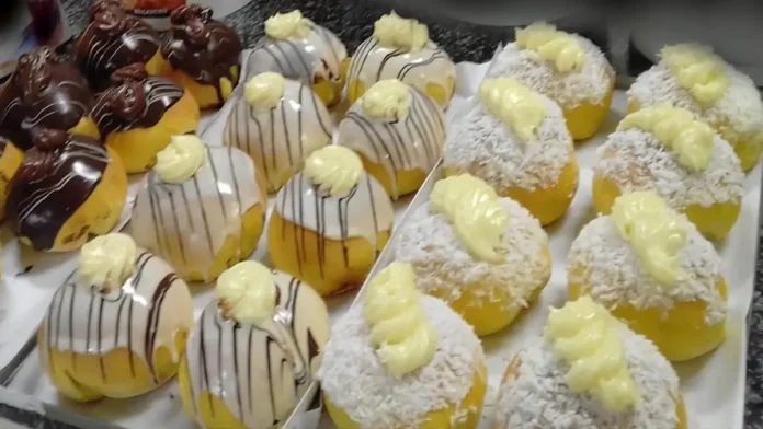 Três bandejas brancas com diferentes tipos de pães. A primeira bandeja contém pães redondos cobertos com coco e creme. A segunda bandeja tem pães cobertos com ganache branco, riscos de chocolate preto e gotas de creme. A terceira bandeja apresenta pães cobertos com chocolate preto e gotas de creme de chocolate.