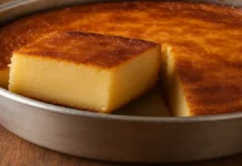 Bolo Mole de Liquidificador: Uma Delícia Simples e Irresistível A imagem mostra um bolo mole recém-assado, com uma crosta dourada e brilhante, e um pedaço cortado revelando seu interior cremoso e macio, semelhante a um pudim. O bolo está disposto em uma forma metálica simples, com uma textura suave e homogênea. O fundo é uma mesa de madeira rústica, criando uma atmosfera acolhedora e caseira.