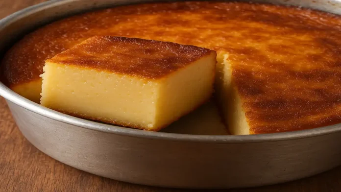 A imagem mostra um bolo mole recém-assado, com uma crosta dourada e brilhante, e um pedaço cortado revelando seu interior cremoso e macio, semelhante a um pudim. O bolo está disposto em uma forma metálica simples, com uma textura suave e homogênea. O fundo é uma mesa de madeira rústica, criando uma atmosfera acolhedora e caseira.