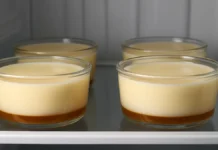 Pudim que não vai ao forno. Pequenos potes de vidro transparentes, alinhados em uma prateleira de geladeira, contêm pudins gelados ainda intocados. A base de cada pudim tem uma camada dourada de caramelo, enquanto a parte superior exibe um creme claro e uniforme. A luz fria da geladeira ilumina suavemente a cena, destacando a textura lisa da sobremesa.